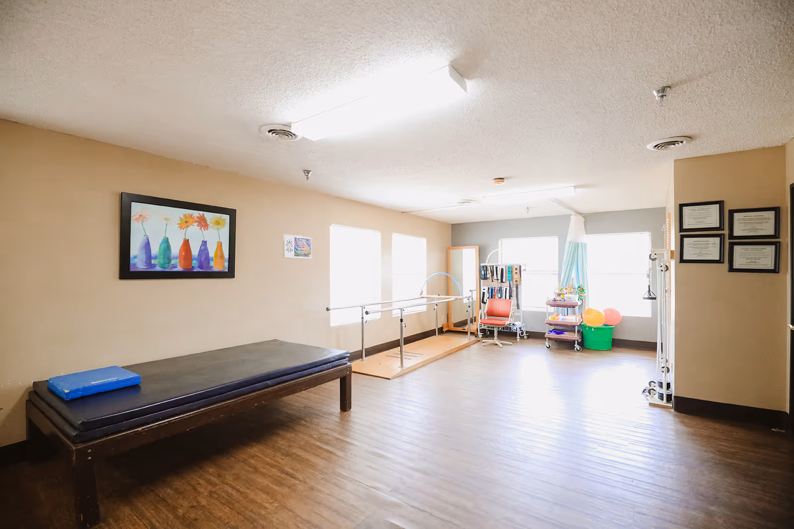 A bright rehabilitation room with wooden flooring, a padded therapy table with a blue cushion, parallel bars for walking exercises, a red chair, a full-length mirror, and various therapy equipment including colorful balls and resistance bands. The walls are beige with framed artwork and certificates.
