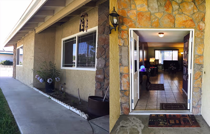 View of the exterior side of a building with a concrete walkway, a window, and a small garden bed with plants. Next to it is an open doorway with double doors leading into a warmly lit interior room with tiled floors, furniture, and lamps visible inside.