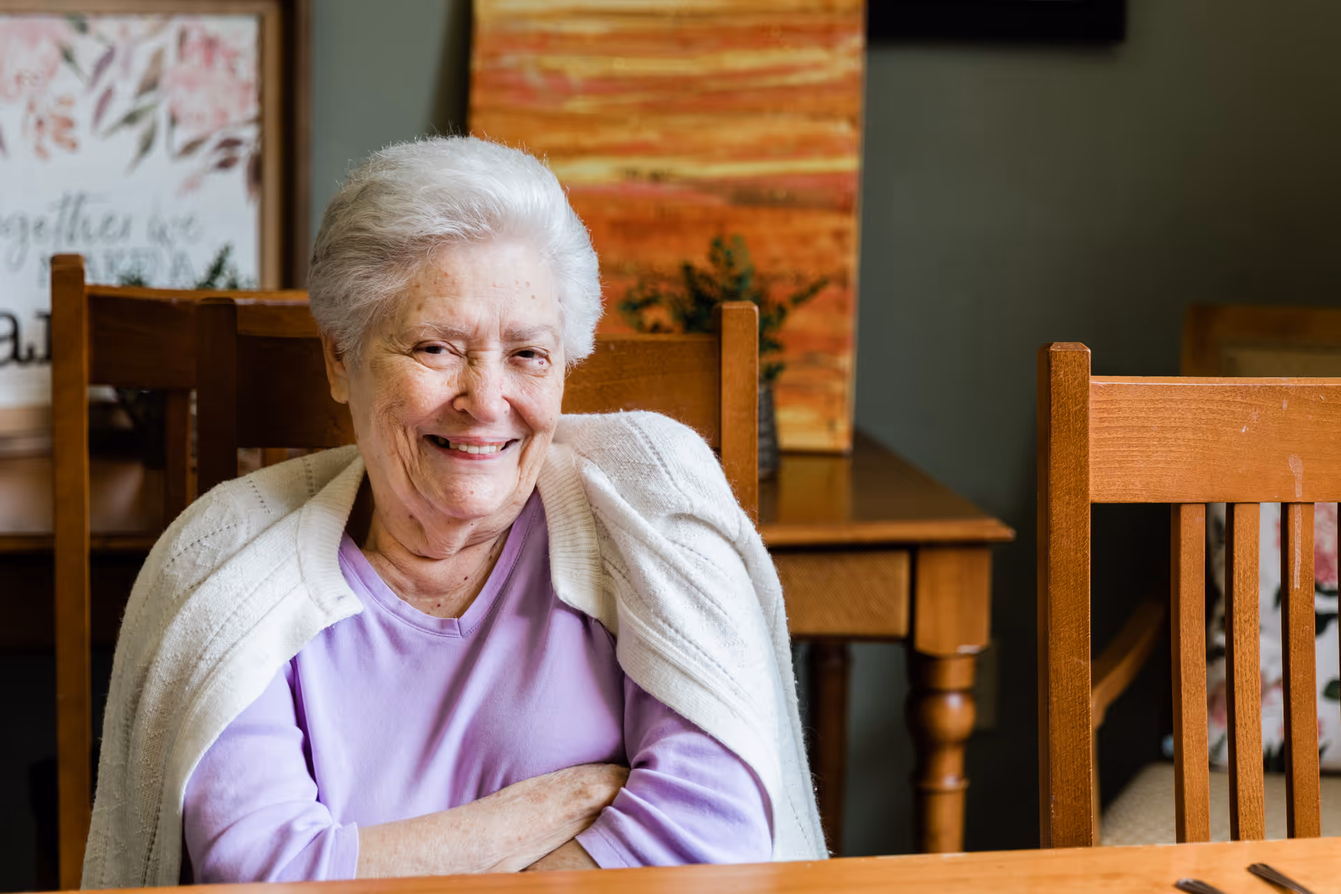 An elderly woman with white hair wearing a lavender shirt and a white cardigan draped over her shoulders is sitting at a wooden table, smiling warmly. Behind her, there is a wooden chair, a table with a small plant, and colorful artwork on the wall.