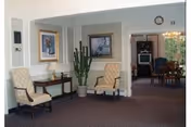 Small seating area in a senior facility with two upholstered chairs beside a side table, a tall potted cactus, framed artwork on the wall, and an open doorway to a dining area.