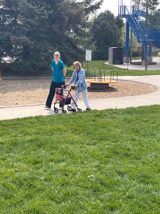 An elderly woman using a walker is accompanied by a caregiver in teal scrubs walking beside her on a paved path in a park-like outdoor area with green grass, trees, and playground equipment in the background.