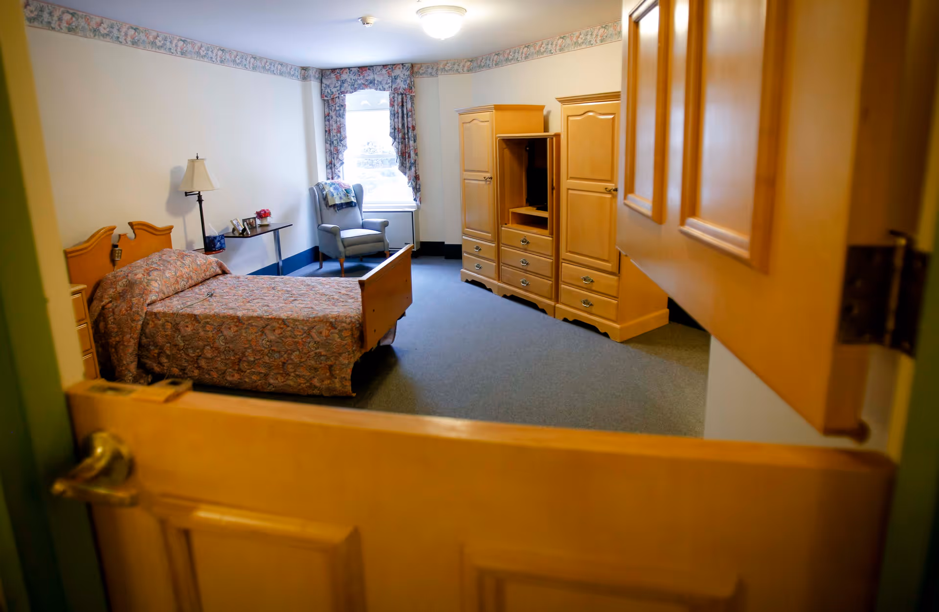 View into a bedroom with a wooden bed covered with a patterned bedspread, a wooden dresser and wardrobe, a small table with a lamp and picture frames, and a cushioned armchair near a window with floral curtains.