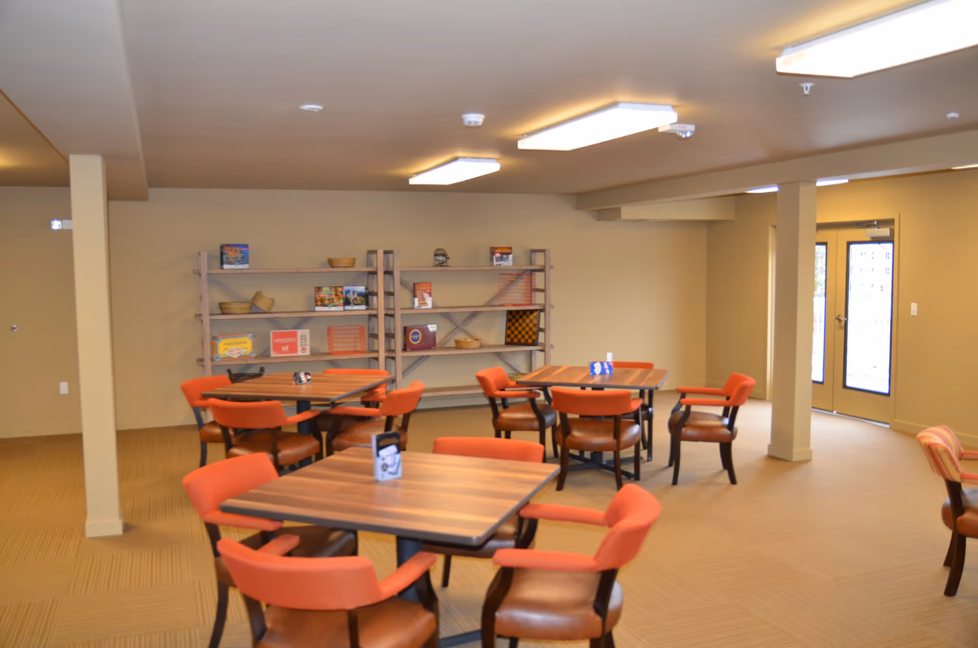 A common area with several square wooden tables and orange cushioned chairs arranged around them. In the background, there are two wooden shelves with board games and baskets. The room has beige walls and carpet, with fluorescent ceiling lights and a double glass door letting in natural light.