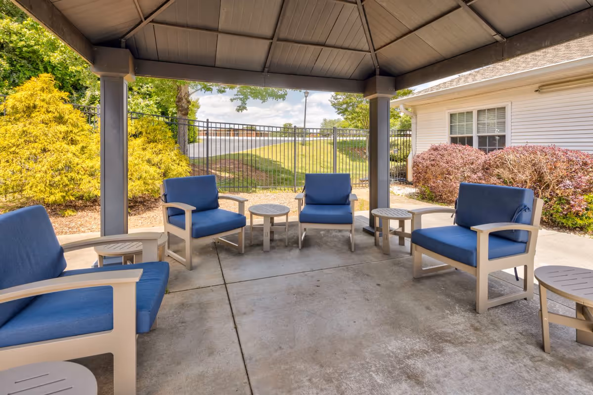 Outdoor covered seating area with blue cushioned chairs and small round tables arranged on a concrete floor, surrounded by greenery and a black metal fence.