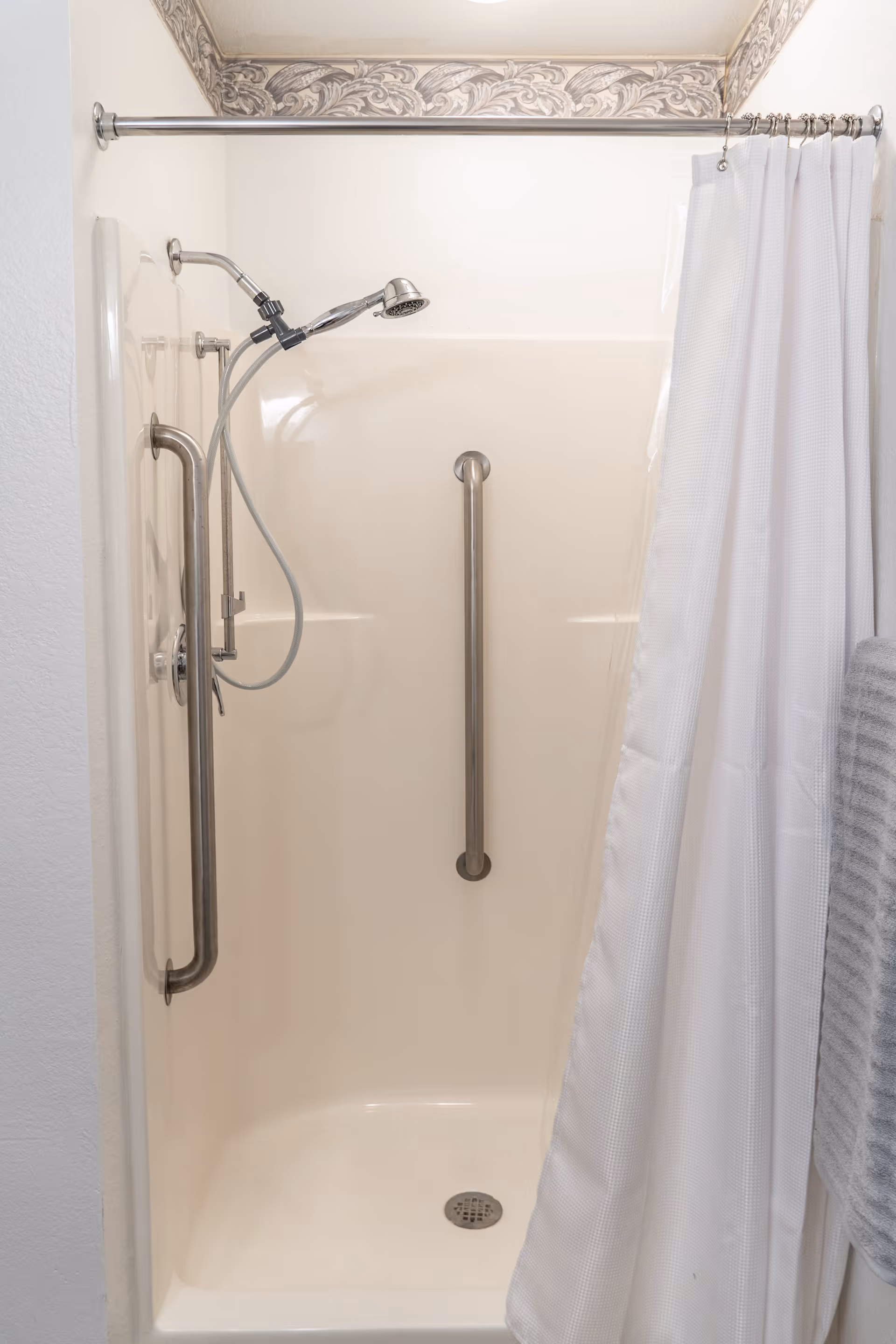 A clean, beige shower stall with two stainless steel grab bars, a handheld showerhead attached to a wall mount, and a white shower curtain partially drawn to the side. There is decorative wallpaper border at the top edge of the wall.