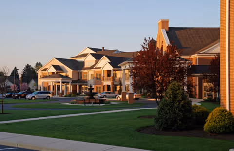 Exterior view of a senior living facility building with multiple peaked roofs, a covered entrance, parked cars, green lawns, trees with autumn foliage, and a fountain in front during sunset.