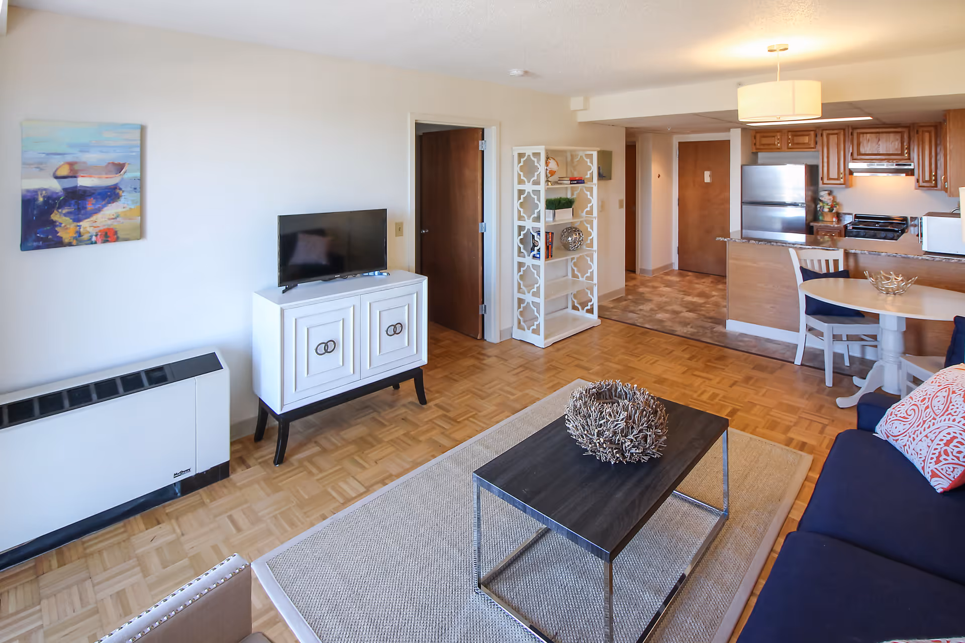 A bright and cozy senior living apartment interior featuring a living room with a navy blue couch adorned with patterned pillows, a dark wood coffee table with a decorative centerpiece, and a white cabinet with a flat-screen TV on top. The room opens into a kitchen area with wooden cabinets, a stainless steel refrigerator, stove, and microwave. A small round dining table with chairs is positioned near the kitchen. The walls are light-colored, and there is a colorful painting of a boat on the wall. The floor is parquet wood, and a beige rug is placed under the coffee table.