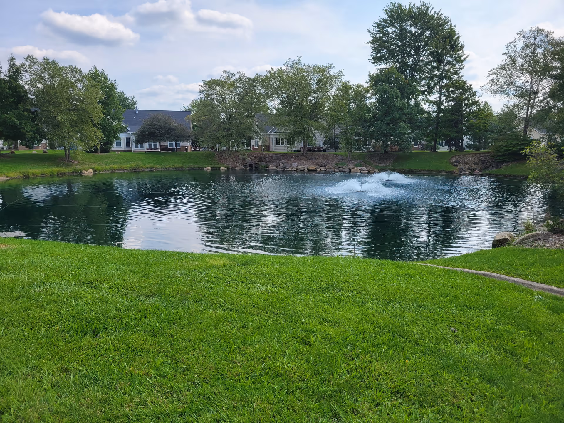A serene outdoor scene featuring a small pond with a water fountain in the center, surrounded by green grass and trees. Residential buildings are visible in the background under a partly cloudy sky.
