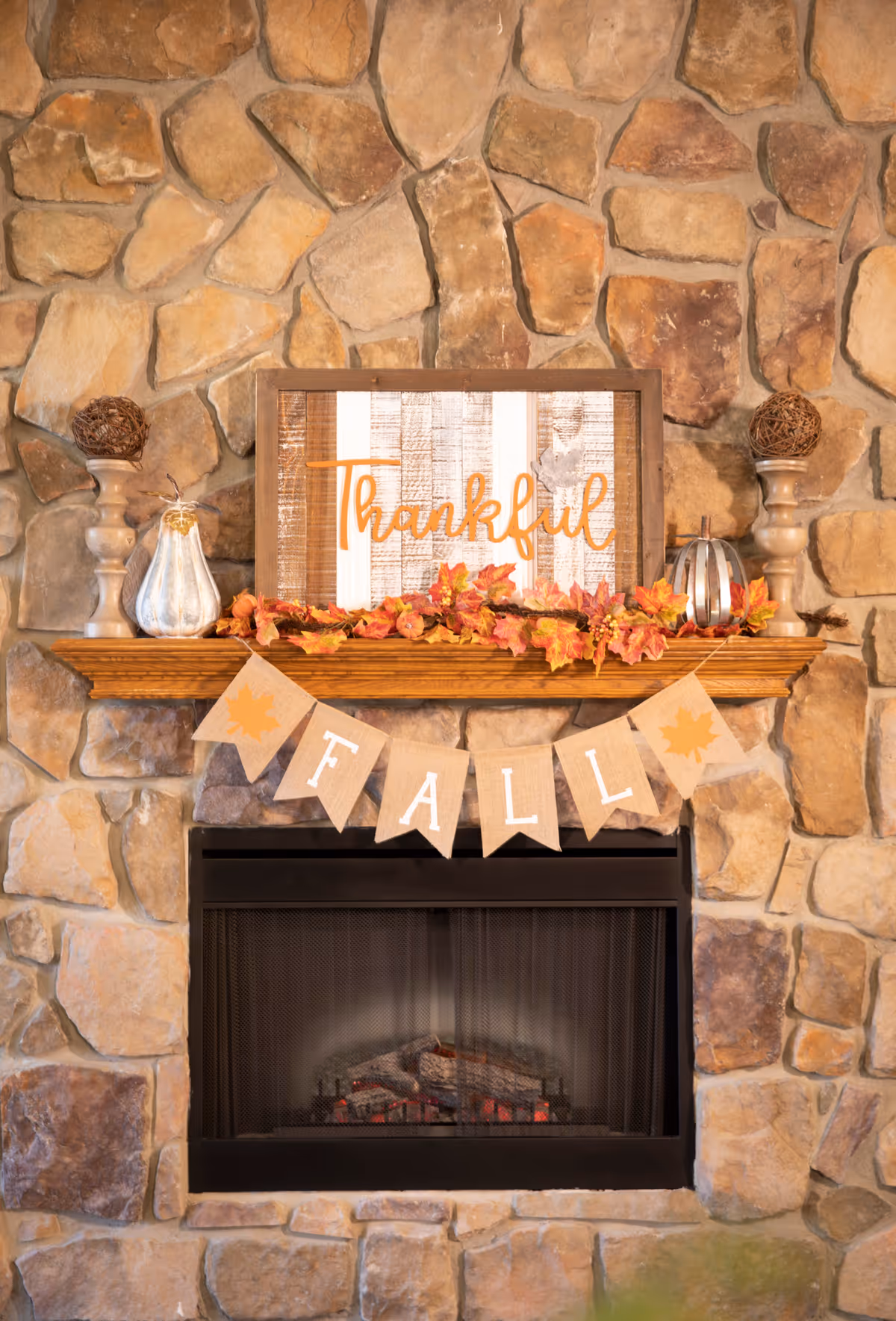 Stone fireplace with a wooden mantel decorated with a 'Thankful' sign, autumn leaves, pumpkins, and a 'FALL' banner.
