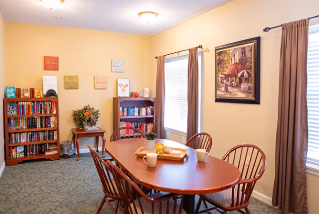 A cozy room with a wooden oval dining table surrounded by six wooden chairs. On the table, there are two white mugs, a tray with a small plant, and some decorative items. The room has beige walls with framed artwork and decorative plaques. There are two windows with brown curtains letting in natural light. Two wooden bookshelves filled with books and a small table with a plant are against the walls.