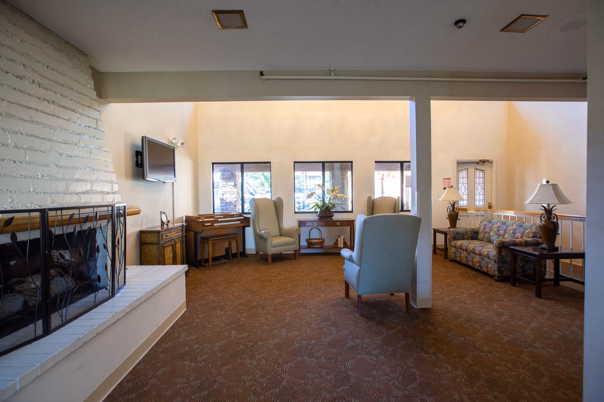 A cozy senior living facility common area with a white brick fireplace on the left, a wall-mounted TV above a wooden cabinet, two beige armchairs, a wooden piano, a floral-patterned sofa, two side tables with lamps, and windows letting in natural light.