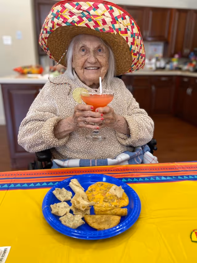 An elderly woman wearing a colorful sombrero and a beige fleece jacket is sitting at a table with a yellow and orange tablecloth. She is holding a glass with a red drink garnished with a lime slice and a straw, smiling at the camera. In front of her on the table is a blue plate with various Mexican-style snacks including chips and cheese dip. The background shows a kitchen area with wooden cabinets.