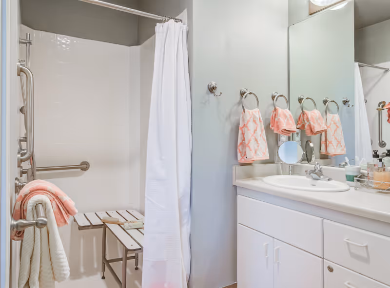 A bathroom with a white shower curtain, a foldable shower bench, grab bars, and a sink with a large mirror above it. Several pink patterned hand towels hang on towel rings on the wall next to the sink.