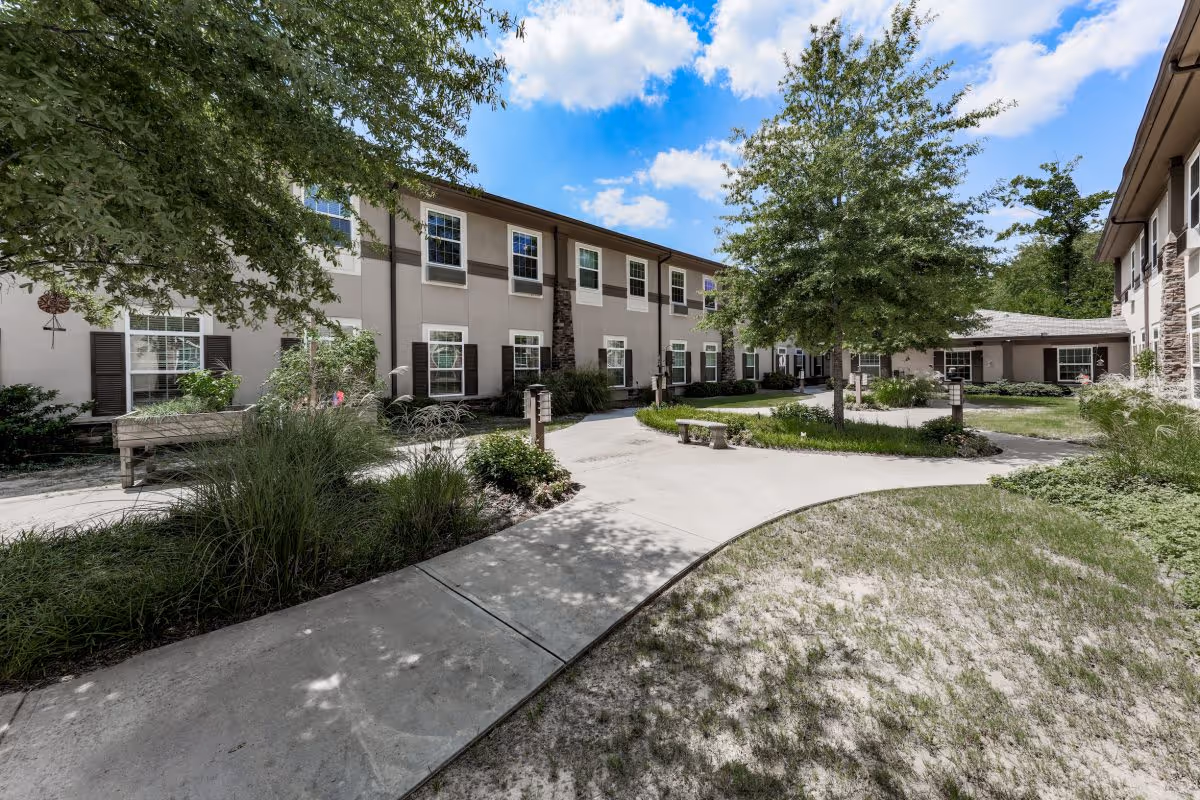 Sunny courtyard with paved walkways, benches, landscaping and a two-story residential building under a blue sky.