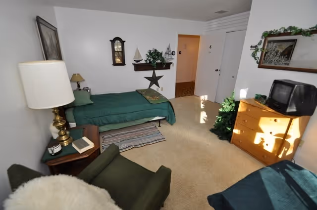 A modest senior living bedroom with a bed, armchair, dresser and small TV.