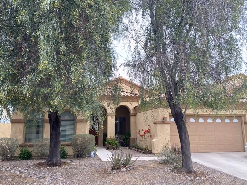 Front exterior of a single-story stucco house with an arched entryway, attached garage, and trees and desert landscaping in the front yard.