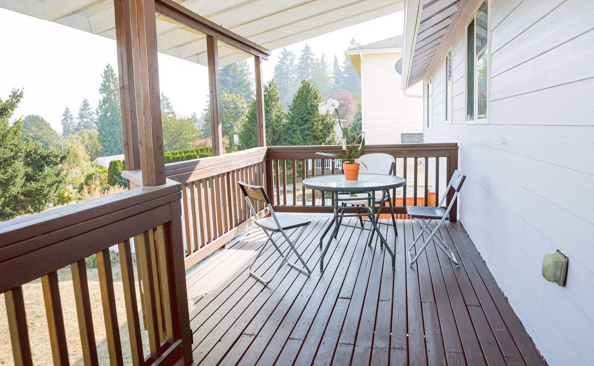 A wooden outdoor deck with a round glass table and three folding chairs. A potted orchid plant is placed on the table. The deck is surrounded by a wooden railing and overlooks a green area with trees and shrubs.