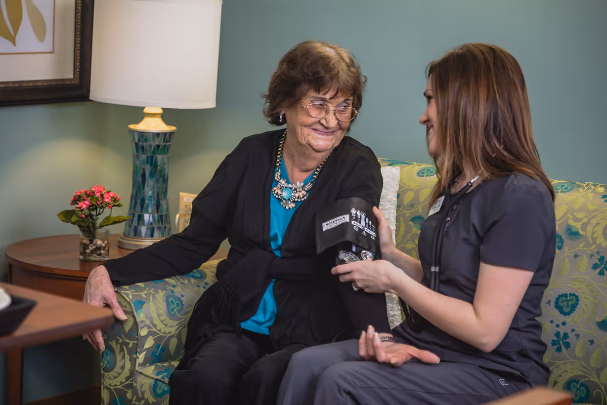 An elderly woman sitting on a patterned couch having her blood pressure taken by a healthcare professional in a cozy room with a lamp and a small flower pot on a side table.