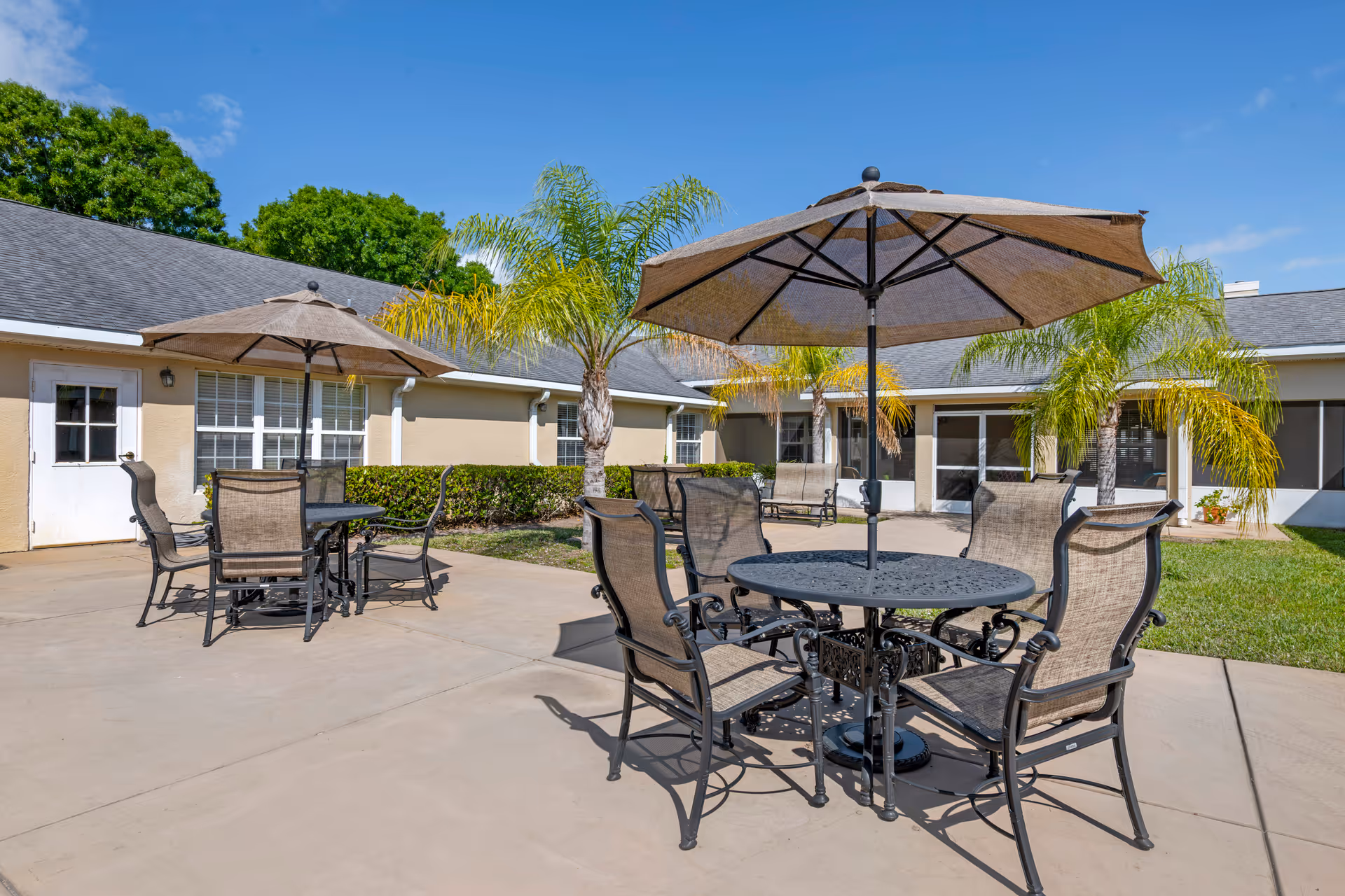 Outdoor patio area with two round metal tables, each surrounded by four mesh chairs and shaded by large umbrellas. The patio is adjacent to a beige building with windows and doors, and there are palm trees and green shrubs around the area under a clear blue sky.