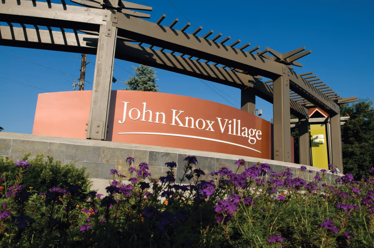 Entrance sign reading 'John Knox Village' mounted on a curved red panel beneath a wooden pergola with purple flowers in the foreground.