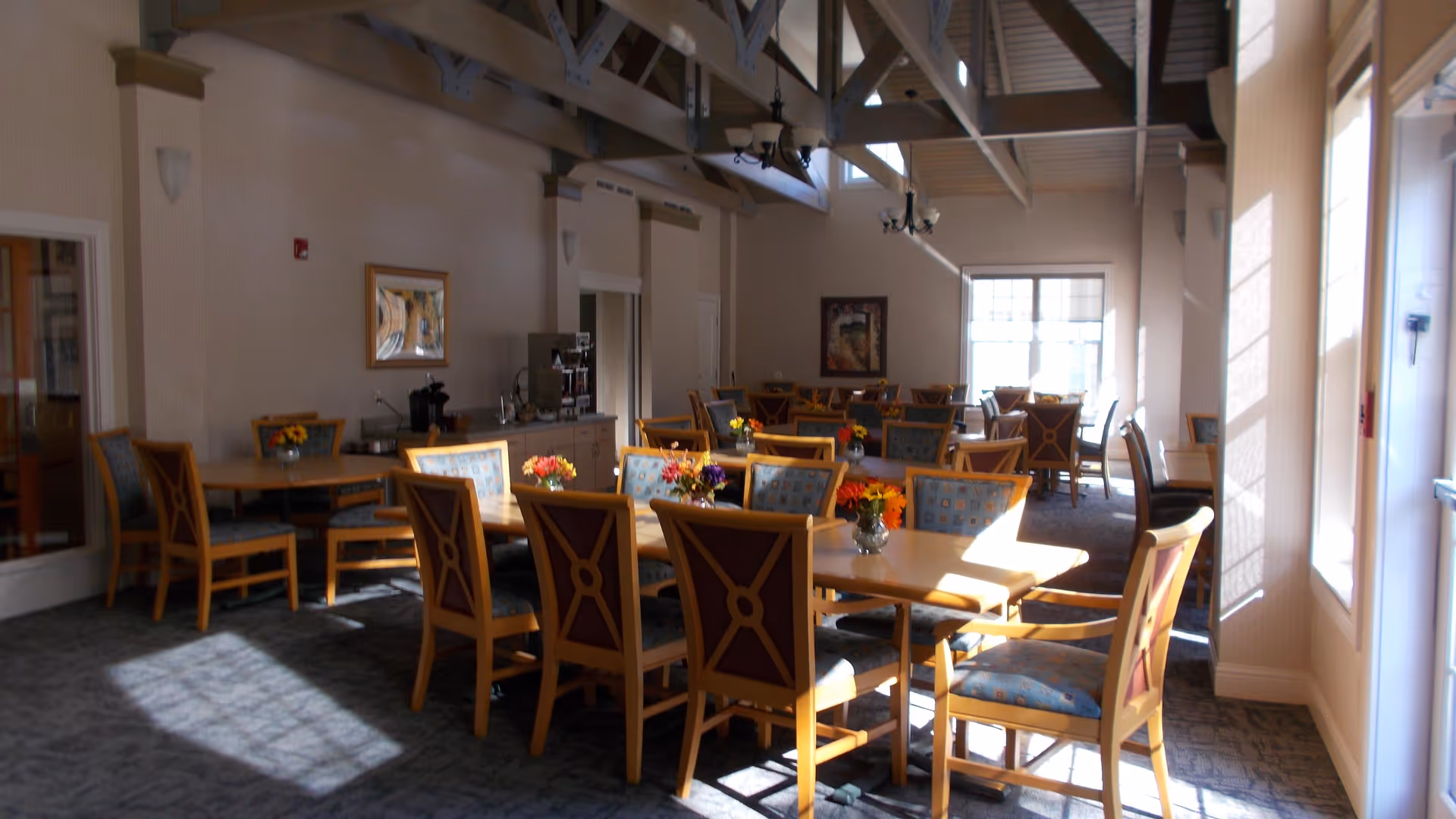 Sunlit communal dining room with wooden tables and chairs, floral centerpieces, and a high beamed ceiling.