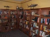 A cozy room with wooden bookshelves filled with books along two walls. The shelves also display decorative items including two fish sculptures and a silver vase. The room has a patterned carpet and beige walls.