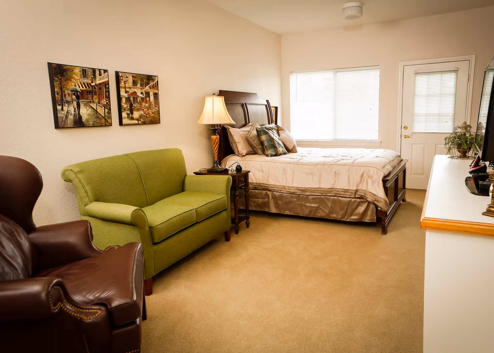 A cozy bedroom in an assisted living facility featuring a bed with beige bedding, a green loveseat, a brown leather armchair, a wooden nightstand with a lamp, two paintings on the wall, a window with blinds, a door, and a white dresser with a TV and plants on top.