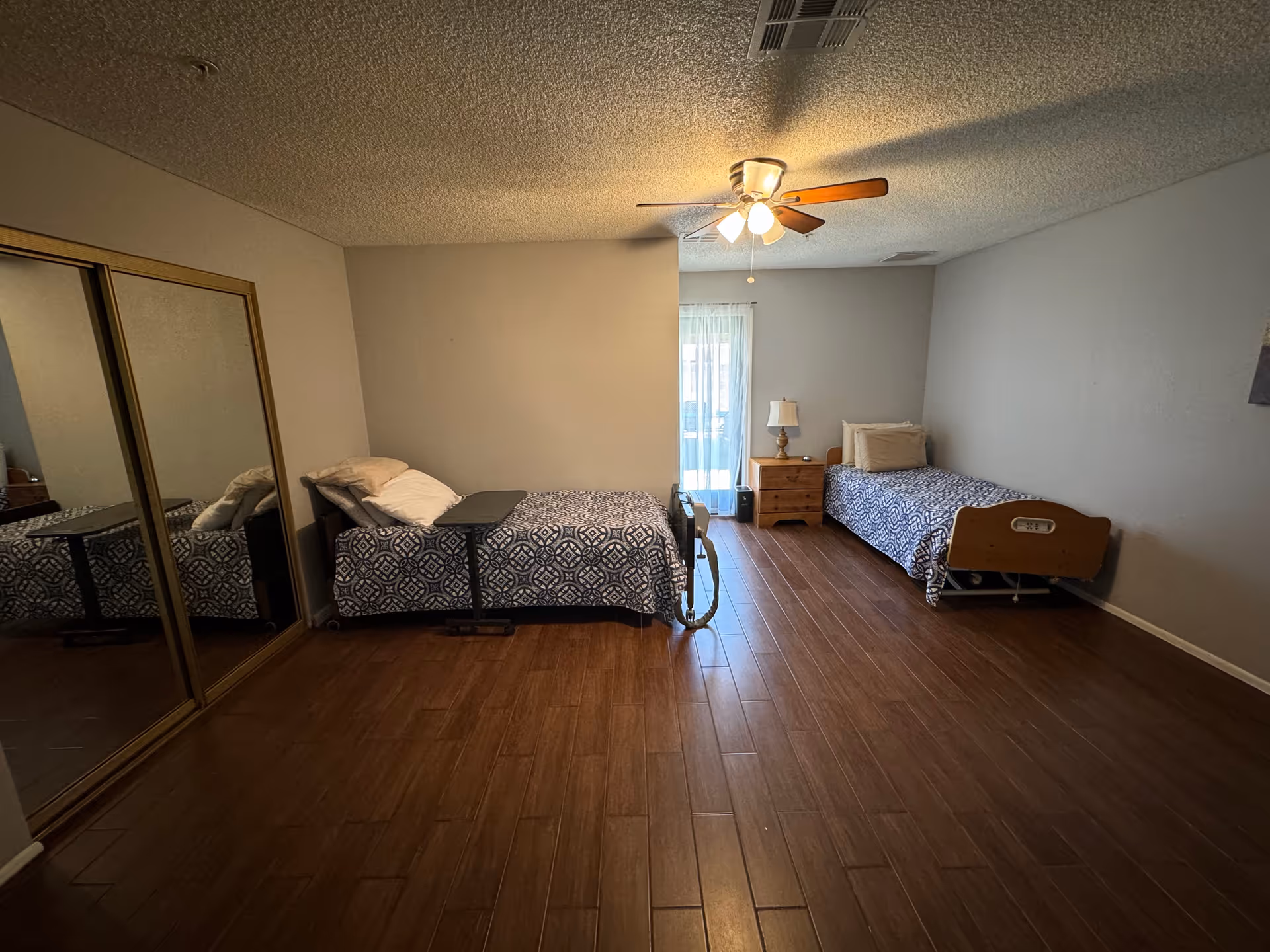 A simple bedroom with two single beds placed against opposite walls. Each bed has a patterned blue and white bedspread and pillows. There is a wooden nightstand with a lamp between the beds near a window with sheer curtains. The room has wooden flooring, a ceiling fan with lights, and a large mirrored closet door on one side.