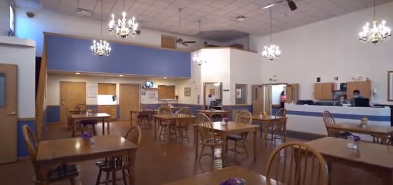 Interior view of a dining room in Victorian Assisted Living facility with wooden tables and chairs arranged neatly. The room has high ceilings with chandeliers, light-colored walls, and a reception desk area where two staff members are seated. There are doors and windows along the walls, and small flower vases are placed on the tables.
