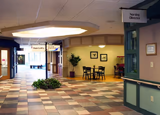 Bright interior hallway of a senior living facility with a tiled floor, a small seating area, plants, and hanging signs including 'Nursing Director'.