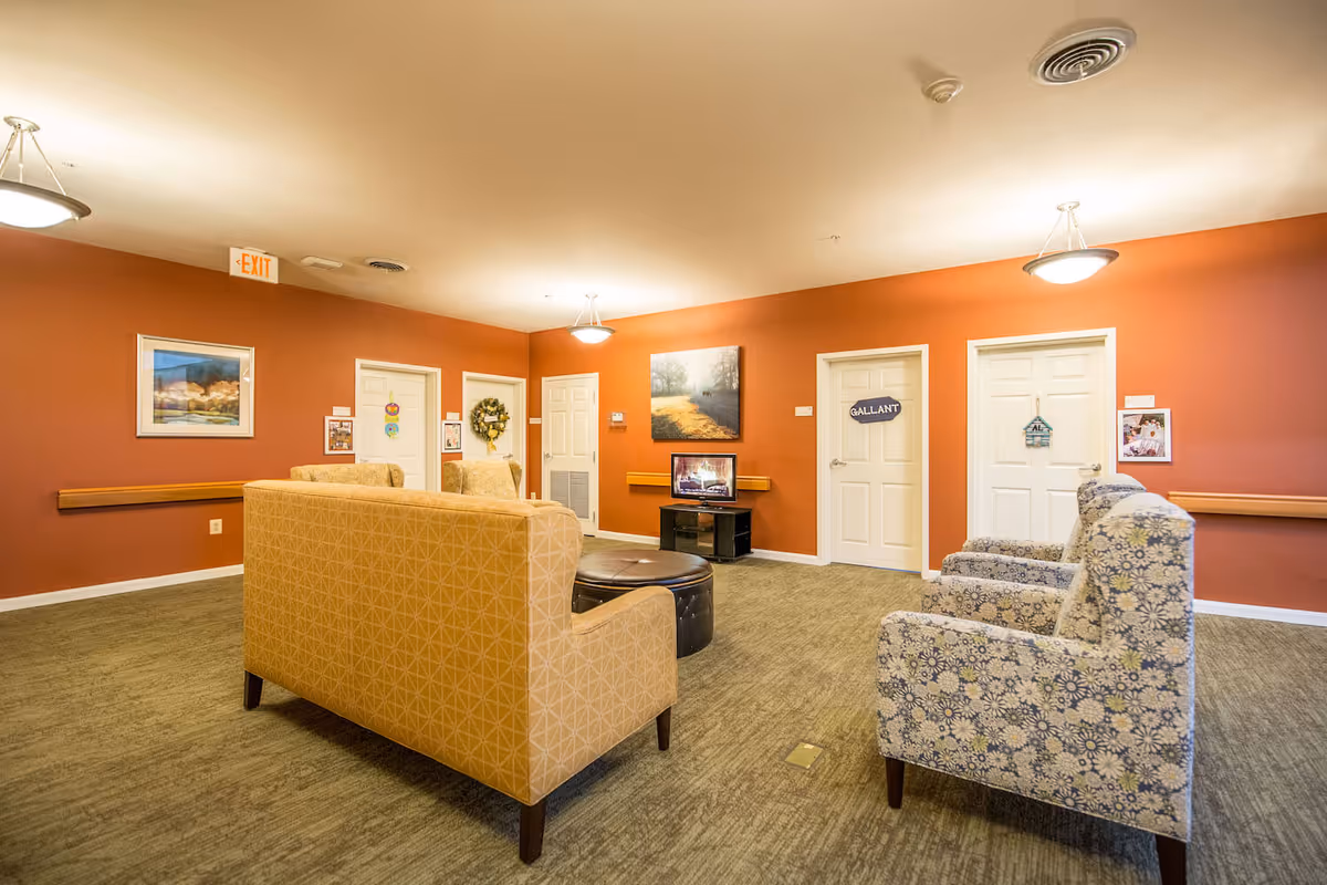 A cozy common area in Arbor Terrace Sudley Manor with orange walls, carpeted floor, and multiple seating options including a patterned yellow sofa and floral armchairs. There is a small TV on a stand displaying a fireplace scene, wall art, and several white doors with name signs. Ceiling lights illuminate the room.
