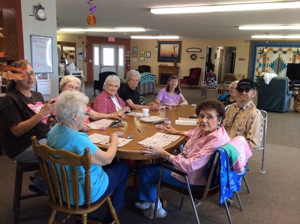 A group of elderly people sitting around a large round table in a common room, playing bingo. The room has comfortable chairs, a quilt hanging on the wall, and a whiteboard with the day's activities listed. The atmosphere appears social and engaging.