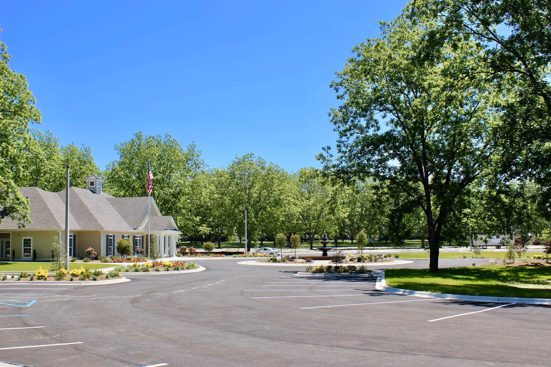 A parking lot with marked parking spaces in front of a single-story building with a pitched roof and an American flag on a flagpole. The area is surrounded by green trees and landscaping under a clear blue sky.