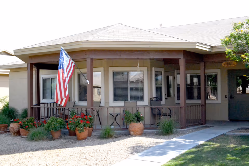 Front porch of a senior living building with an American flag, potted flowers, and seating.