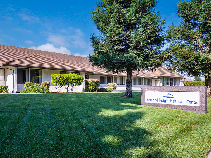 Exterior view of Diamond Ridge Healthcare Center showing a single-story building with a brown roof, multiple windows, neatly trimmed bushes, green lawn, and two large trees under a blue sky with some clouds.