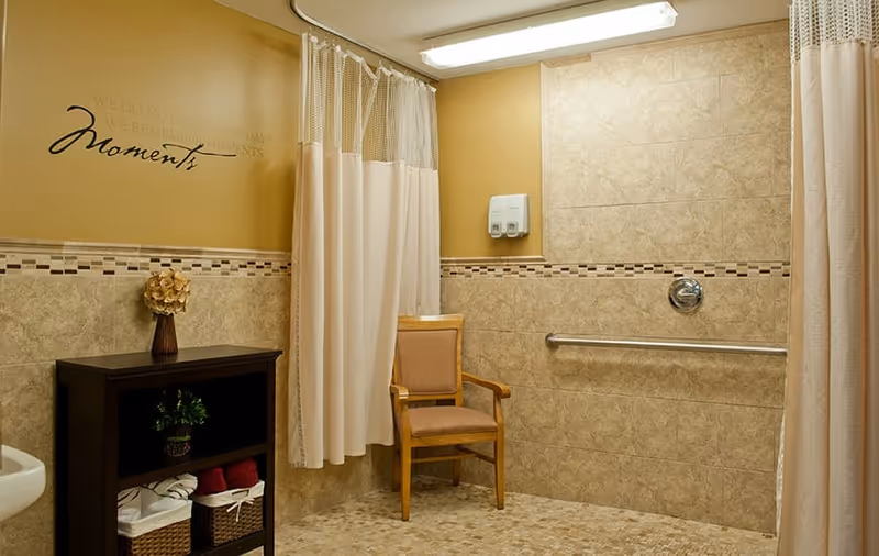 A spacious bathroom with beige tiled walls and floor, featuring a wooden chair, a dark wooden shelf with towels and a plant, a shower area with a grab bar and a soap dispenser, and a curtain for privacy. The wall has a decorative quote that reads 'We do not remember days, we remember moments.'