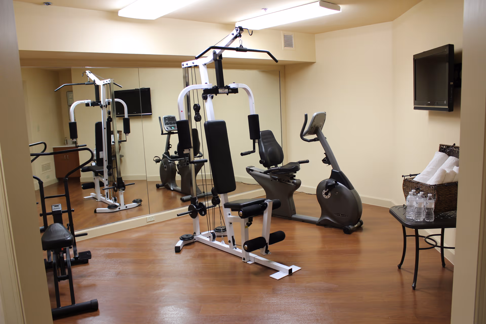 Small fitness room with exercise equipment including a multi-gym machine, a recumbent stationary bike, and a rowing machine. There is a wall-mounted TV, a table with bottled water and a basket of rolled towels, and a large mirror covering one wall. The room has wood flooring and beige walls.