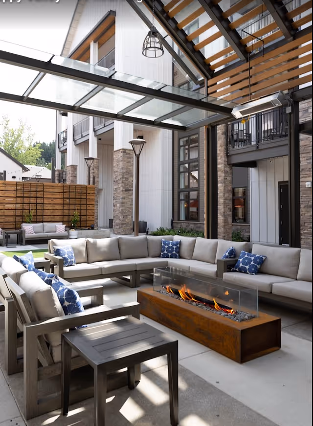 Outdoor seating area at The Springs at Happy Valley featuring a large L-shaped cushioned sofa with blue patterned pillows, wooden chairs, a modern rectangular fire pit with flames, and a glass canopy overhead. The area is surrounded by a building with stone and white panel exterior walls and large windows.