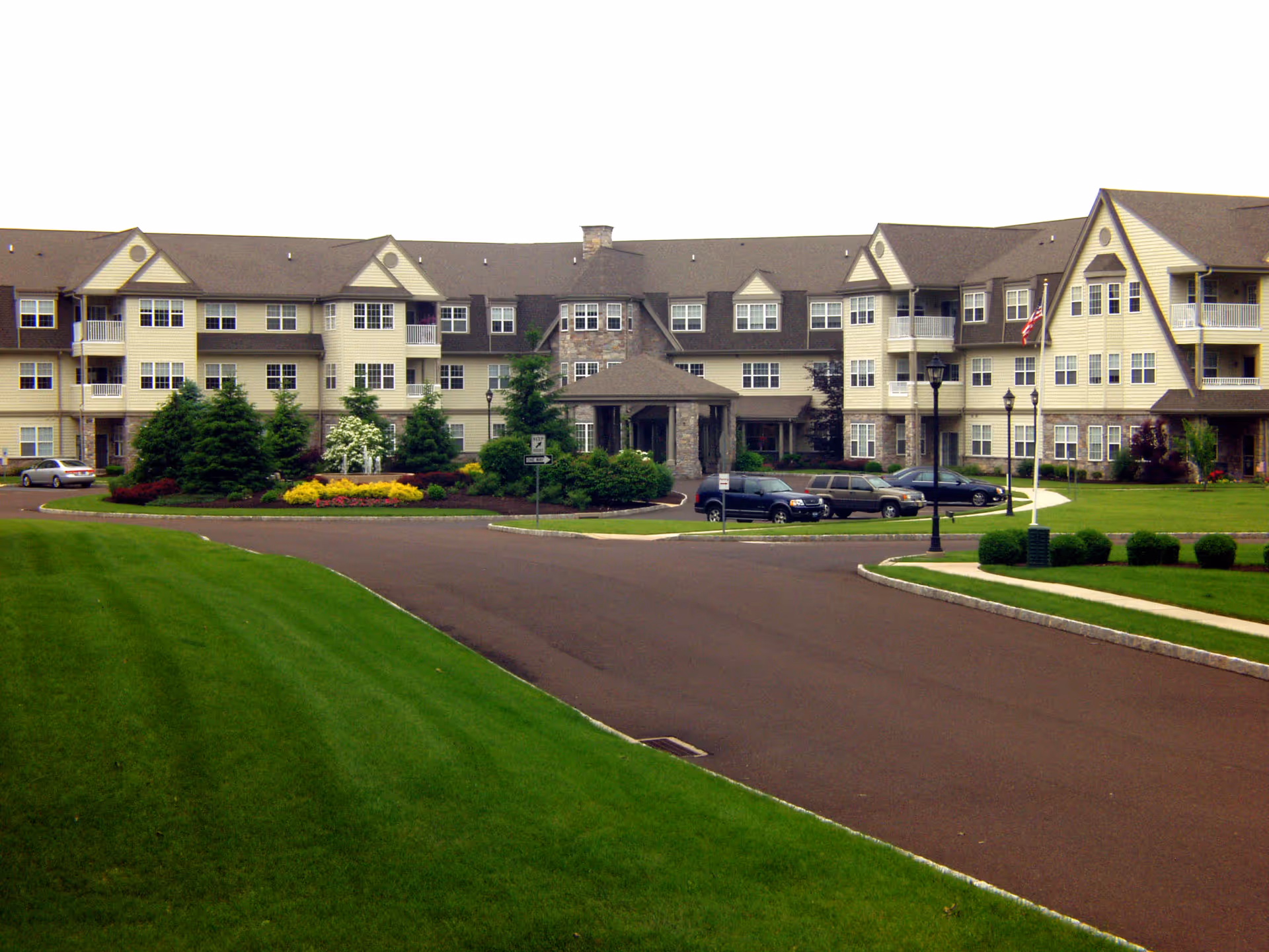 Front exterior of a three-story residential building with balconies, landscaped grounds, a circular driveway, and parked cars.