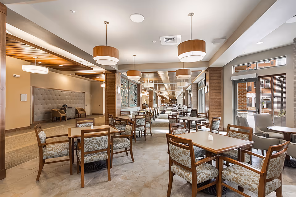A spacious and well-lit dining area in a senior living facility with multiple wooden tables and cushioned chairs arranged neatly. The room features modern pendant lights hanging from the ceiling, large windows allowing natural light to enter, and a decorative wall panel. There are additional seating areas visible in the background.