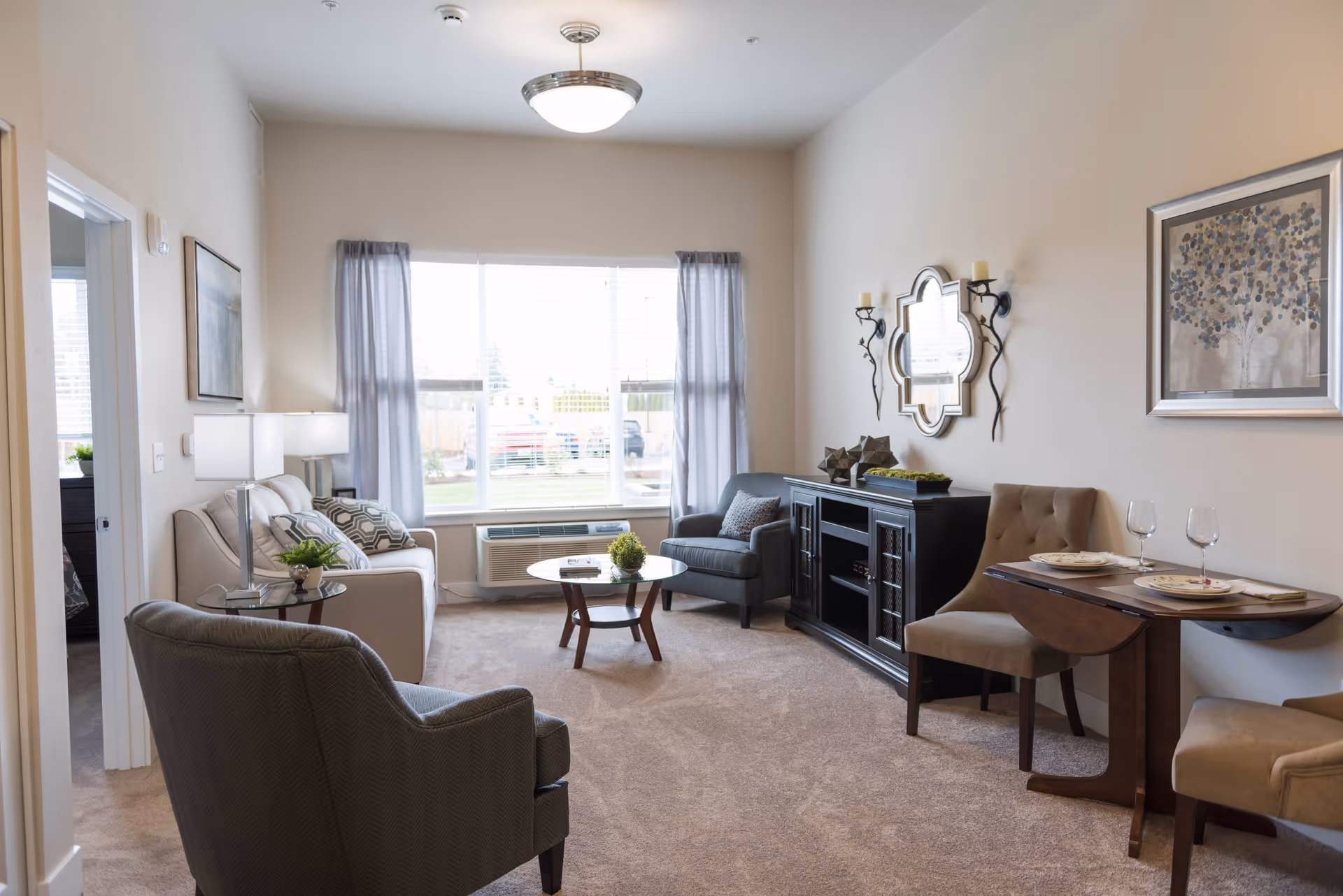 Well-lit living room with seating, coffee table, media console and a small dining nook by a large window.