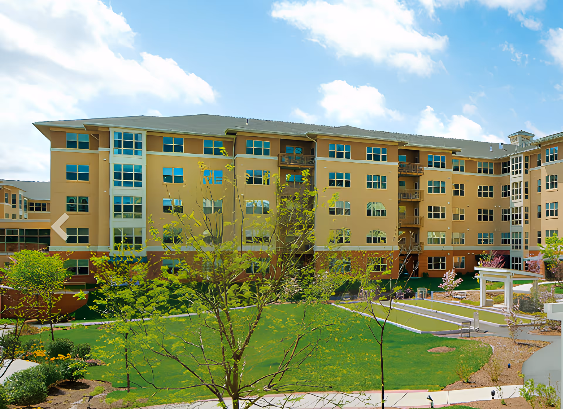 Exterior view of a multi-story senior living facility building with numerous windows and balconies, surrounded by a well-maintained green lawn, trees, and a bocce ball court under a partly cloudy blue sky.