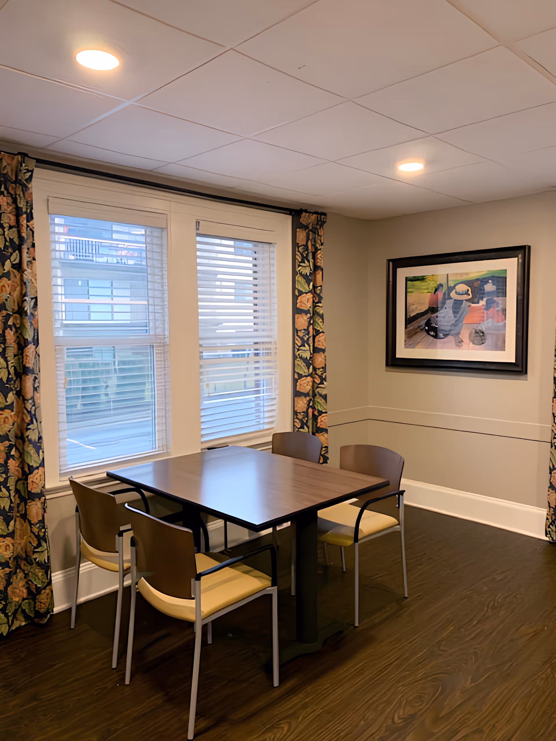 A small dining area with a square wooden table and four chairs with yellow seats and brown backs. The room has two large windows with white blinds and floral curtains. A framed painting hangs on the beige wall to the right. The floor is dark wood, and the ceiling has recessed lighting.
