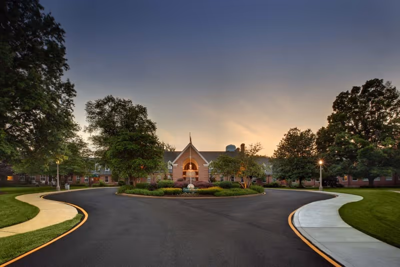 Wide driveway leading to the front entrance of a large brick building surrounded by trees and landscaped greenery at sunset.