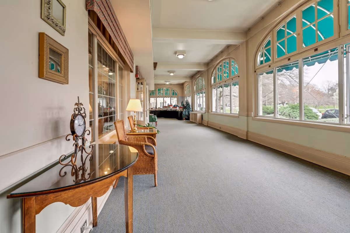 A long, carpeted hallway with large arched windows on the right side letting in natural light. The windows have green awnings outside. On the left side, there is a wooden table with a decorative clock and two wicker chairs with a lamp between them. At the far end of the hallway, there is a Christmas tree and some furniture.