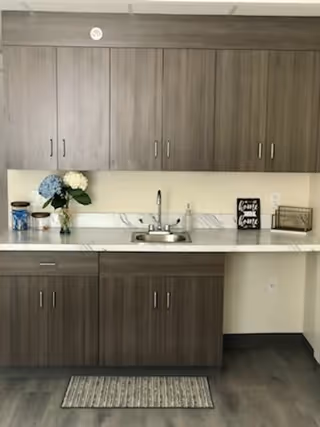 A kitchenette area with wooden cabinets above and below a marble countertop. The countertop features a small sink with a faucet, a vase with blue and white flowers, a container with utensils, a small framed sign, and a metal basket. There is a small rug on the floor in front of the cabinets.