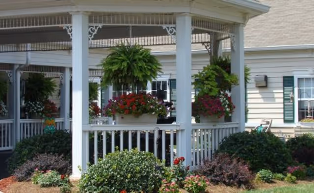 White gazebo-style porch with hanging ferns and colorful flower boxes in front of a beige residential building.