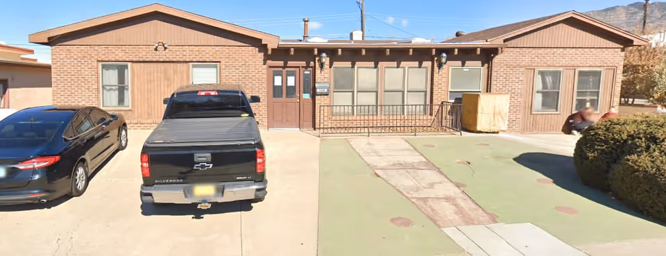 Front exterior view of a single-story brick building with two parked vehicles in front, a black pickup truck and a black sedan. The building has multiple windows and two main doors, with a small ramp leading to the entrance. There are bushes on the right side and mountains visible in the background under a clear blue sky.