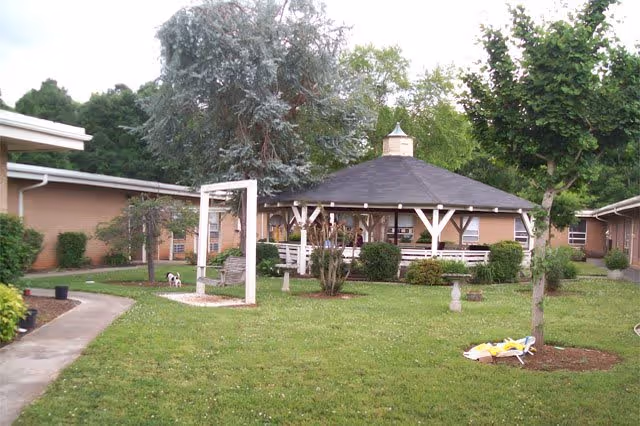 Outdoor courtyard area with a large gazebo surrounded by greenery, trees, and bushes. There is a swing set on the left side and a paved walkway curving through the grass. The buildings of the facility are visible in the background.