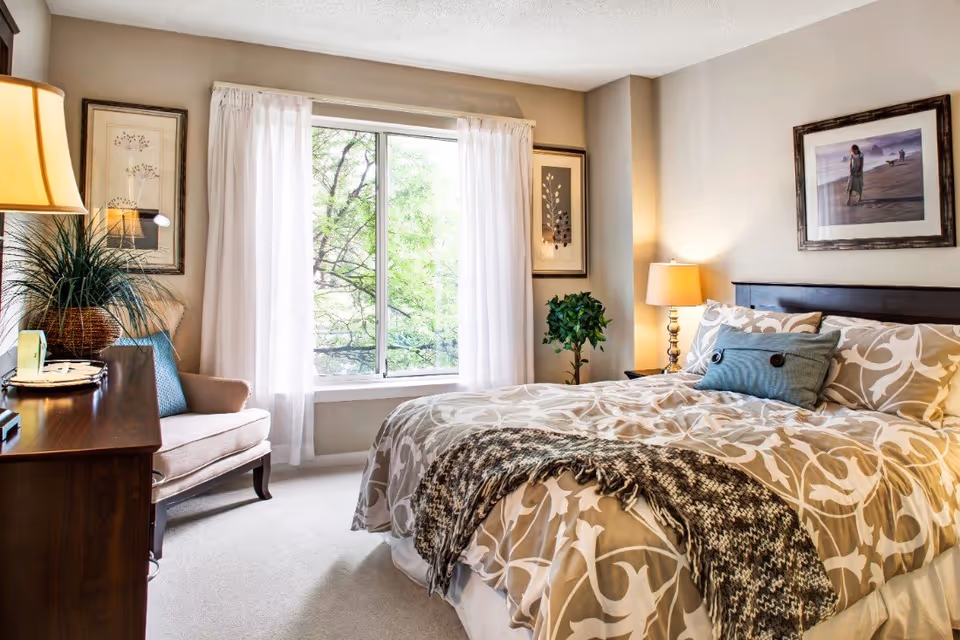 A cozy bedroom with a large bed covered in beige and white patterned bedding, a blue decorative pillow, and a knitted throw blanket. There is a wooden nightstand with a lamp on the right side of the bed, a framed picture hanging above the headboard, and a large window with white curtains letting in natural light. On the left side, there is a wooden dresser with a lamp and a potted plant, and a cushioned armchair next to it. The walls are light-colored, and the room has a calm and inviting atmosphere.
